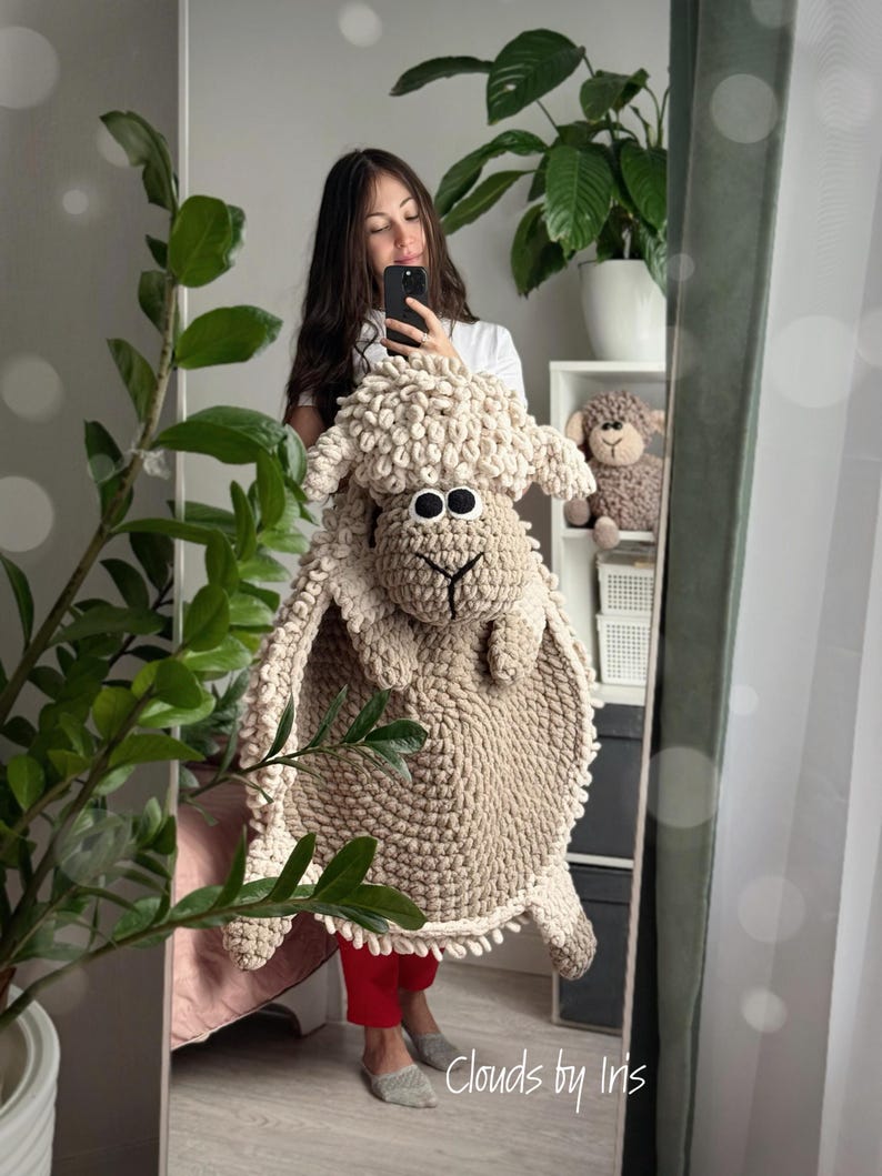 May include: A beige crocheted sheep rug with a fluffy texture. The rug has a large head with black eyes and a brown nose. The rug is being held by a person in a white shirt and red pants. The text "Clouds by Iris" is visible in the bottom right corner of the image.