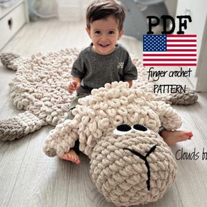 Puede incluir: Una alfombra de crochet con forma de oveja con una textura esponjosa. La alfombra es beige y tiene una nariz marrón y ojos negros. La alfombra está colocada sobre un suelo de madera. Un niño está sentado en la alfombra. La imagen incluye el texto "PDF finger crochet PATTERN Clouds by Iris".