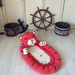 Peut inclure: Un nid de bébé en crochet en forme de tigre rose et blanc avec des accents noirs. Le nid est en forme de tigre avec un visage endormi et des pattes. Le texte "Clouds by Tris" est visible sur le nid.
