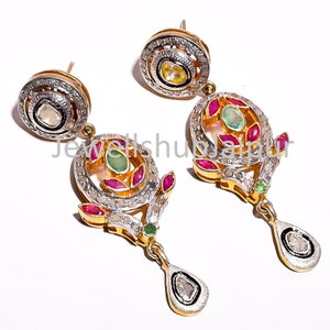 Peut inclure: Boucles d'oreilles dorées avec des détails complexes. Chaque boucle d'oreille présente une pierre précieuse centrale entourée de petites pierres et d'un halo de minuscules pierres. Le design comprend des accents de rubis et d'émeraudes, avec un pendentif en forme de goutte.