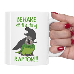 May include: White ceramic mug featuring a green parrot dressed as a dinosaur. The mug has the text "Beware of the tiny RAPTOR!!!" printed on it.