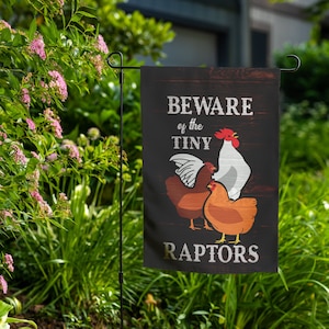 May include: A black garden flag with a white background and the text "Beware of the Tiny Raptors" in white lettering. Three chickens are depicted on the flag, one white and two brown.
