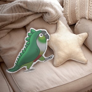 Pu&ograve; includere: Un cuscino a forma di pappagallo verde e rosso con un design a costume di dinosauro &egrave; appoggiato su un divano beige, accanto a un cuscino a forma di stella e a un cuscino a rullo. Una coperta beige lavorata a maglia e cuscini decorativi completano la scena.