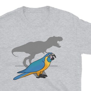 Puede incluir: Camiseta gris claro con un guacamayo de dibujos animados con plumas azules y amarillas, y una sombra gris de un Tyrannosaurus Rex. El guacamayo está sobre una fina línea gris. La camiseta es de material suave.