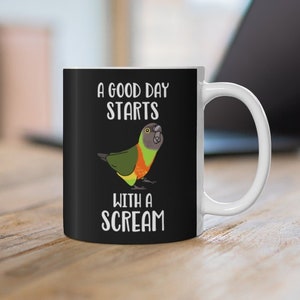 May include: A white ceramic mug with a black design. The design features a parrot with the text "A good day starts with a scream".