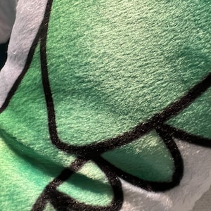 Pu&ograve; includere: Primo piano di un peluche con una superficie verde strutturata e dettagli di contorno neri. Il giocattolo ha un bordo bianco. Il design sembra essere un personaggio dei cartoni animati.