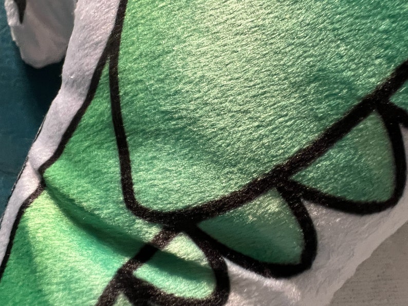 Pu&ograve; includere: Primo piano di un peluche con una superficie verde strutturata e dettagli di contorno neri. Il giocattolo ha un bordo bianco. Il design sembra essere un personaggio dei cartoni animati.