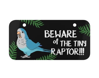 May include: A black sign with a green leafy border and a blue and gray parrot. The sign reads "Beware of the Tiny Raptor!!!"