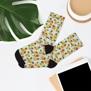 May include: A pair of light blue socks with a repeating pattern of yellow and orange parakeets. The socks are folded in half and laid out on a white surface.
