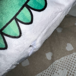 Pu&ograve; includere: Un cuscino di peluche bianco con un disegno di dinosauro verde. I denti del dinosauro sono delineati in nero. Una cerniera bianca &egrave; visibile sul lato. Il cuscino &egrave; su una superficie a fantasia.