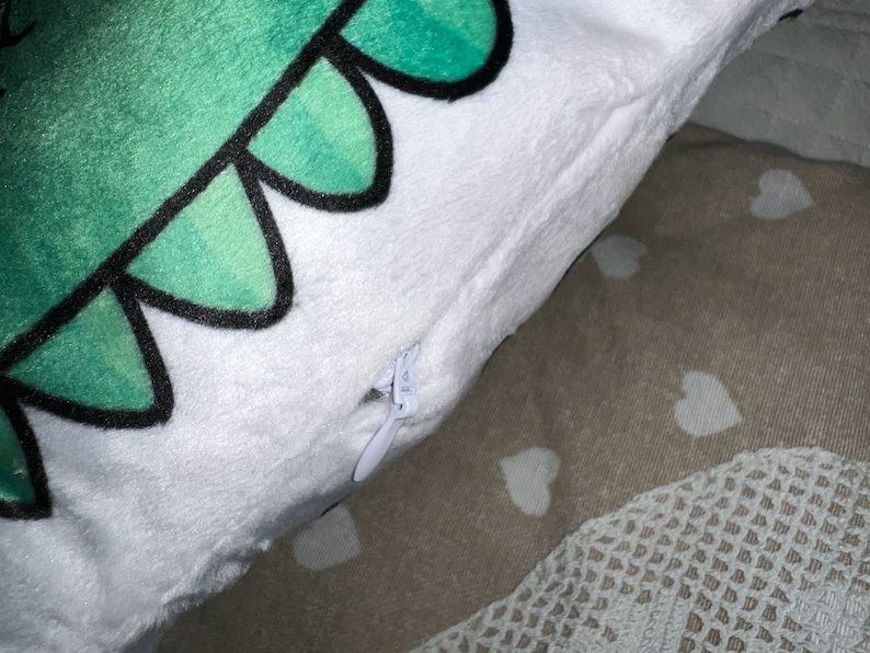 Pu&ograve; includere: Un cuscino di peluche bianco con un disegno di dinosauro verde. I denti del dinosauro sono delineati in nero. Una cerniera bianca &egrave; visibile sul lato. Il cuscino &egrave; su una superficie a fantasia.