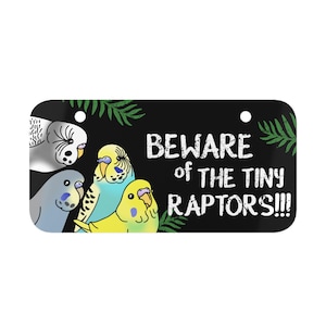 以下が含まれることがあります： 「Beware of the Tiny Raptors!!!」と白文字で書かれた黒い看板。看板には、灰色のオウム、青緑色のオウム、黄色のオウム2羽など、4羽のオウムが描かれている。