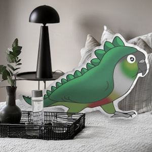 Pu&ograve; includere: Un cuscino decorativo a forma di pappagallo verde con un costume da dinosauro. Il cuscino ha uno sfondo bianco ed &egrave; posto su un letto con cuscini grigi. Il pappagallo ha un corpo verde, un petto rosso e una testa grigia.