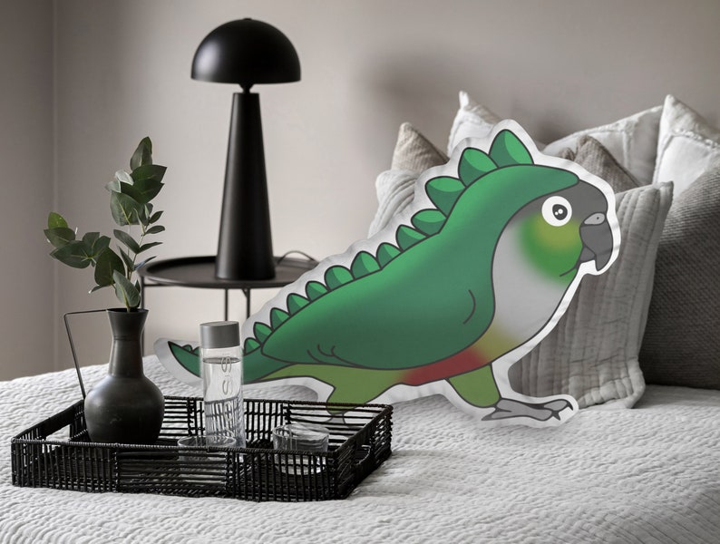 Pu&ograve; includere: Un cuscino decorativo a forma di pappagallo verde con un costume da dinosauro. Il cuscino ha uno sfondo bianco ed &egrave; posto su un letto con cuscini grigi. Il pappagallo ha un corpo verde, un petto rosso e una testa grigia.