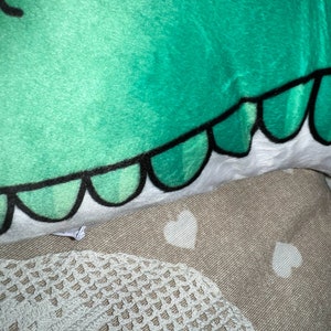 Pu&ograve; includere: Primo piano di una testa di dinosauro di peluche verde con contorni neri e dettagli bianchi. Le squame del dinosauro sono delineate in nero. Il peluche &egrave; appoggiato su una superficie beige con motivi a cuore bianchi e un bordo in pizzo bianco.