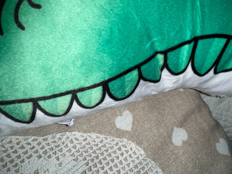 Pu&ograve; includere: Primo piano di una testa di dinosauro di peluche verde con contorni neri e dettagli bianchi. Le squame del dinosauro sono delineate in nero. Il peluche &egrave; appoggiato su una superficie beige con motivi a cuore bianchi e un bordo in pizzo bianco.