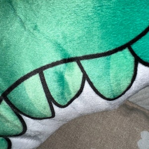 Pu&ograve; includere: Primo piano di un peluche con un design verde e bianco. Il giocattolo ha una superficie morbida e strutturata con un motivo di scaglie verdi bordate di nero. La sezione bianca ha un bordo smerlato.