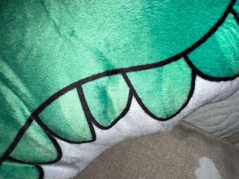 Pu&ograve; includere: Primo piano di un peluche con un design verde e bianco. Il giocattolo ha una superficie morbida e strutturata con un motivo di scaglie verdi bordate di nero. La sezione bianca ha un bordo smerlato.