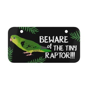 以下が含まれることがあります： 緑の葉と緑と黄色のオウムが描かれた黒い看板。看板には「Beware of the Tiny Raptor!!!」と書かれている