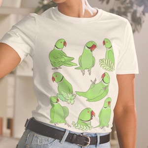 May include: White t-shirt featuring a playful illustration of several green parrots with red beaks. The parrots are in various poses, some perched and others reclining. The design is a whimsical and colorful depiction of the birds.