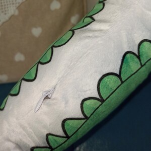 Pu&ograve; includere: Un cuscino di peluche bianco con un bordo smerlato verde, bordato di nero. Una cerniera bianca &egrave; visibile sul lato. Il cuscino &egrave; su una superficie blu.