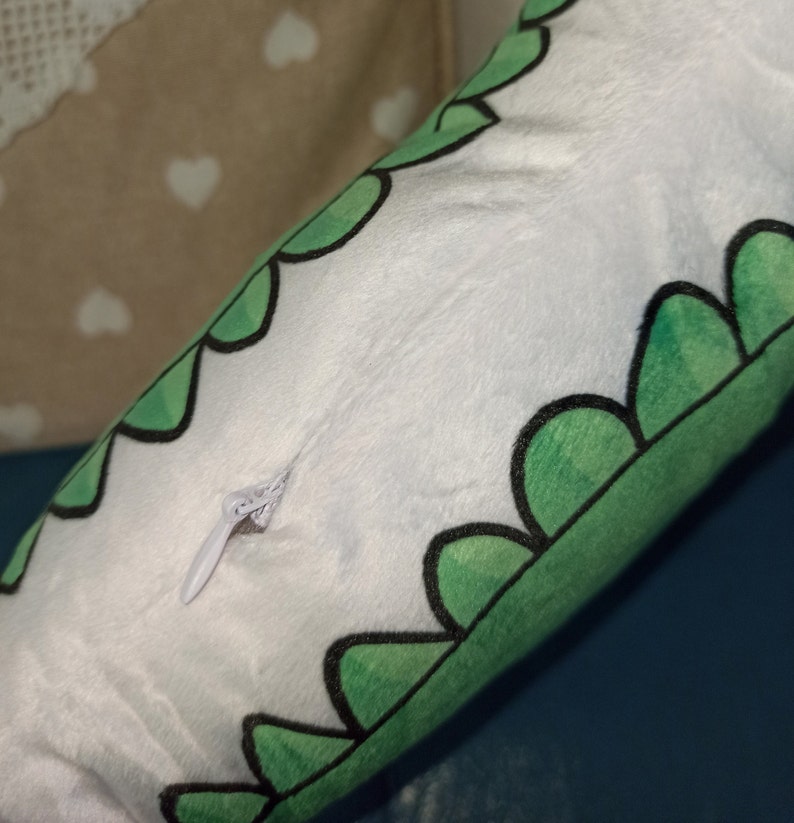 Pu&ograve; includere: Un cuscino di peluche bianco con un bordo smerlato verde, bordato di nero. Una cerniera bianca &egrave; visibile sul lato. Il cuscino &egrave; su una superficie blu.