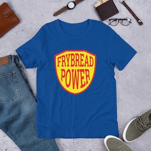 May include: A royal blue t-shirt featuring a yellow and red shield design. The shield displays the text "Frybread Power" in red lettering.