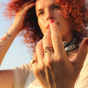 May include: A woman's hand wearing three gold rings with black gemstones. The rings are on the middle finger and the index finger. The woman has red curly hair and is wearing a white shirt.