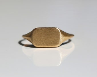 May include: A gold signet ring with a brushed finish. The ring has a rectangular top and a rounded band.