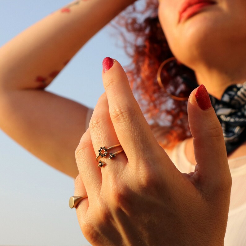 May include: A close-up of a hand wearing a gold ring with black accents. The ring is a delicate design with multiple small flower-like shapes. The hand is also wearing a silver ring on the ring finger.