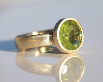 May include: A gold ring featuring a round, faceted, lime-green peridot gemstone set in a smooth bezel setting. The band has a brushed, matte finish, and the entire piece is captured with a clear reflection on a smooth, light-coloured surface.