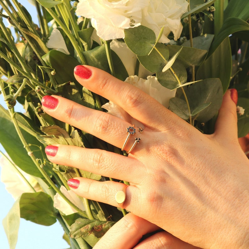 May include: A hand wearing two gold rings. The ring on the index finger has a small flower design with black stones. The ring on the middle finger is a simple oval band. The hand is surrounded by white flowers.