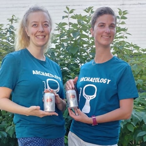Puede incluir: Dos personas con camisetas azul turquesa con la palabra "ARQUEOLOGÍA" impresa en la parte delantera. Las camisetas tienen un contorno blanco de una cerradura. Ambas personas sostienen una lata plateada.