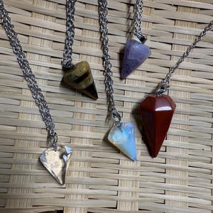 May include: Four necklaces featuring silver-tone metal chains and pointed, faceted gemstone pendants. The pendants include a clear quartz, a brown and black striped tiger's eye, a translucent white opal-like stone, and a solid red jasper stone.