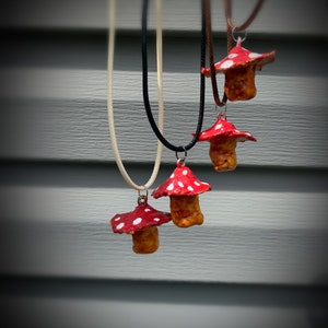 May include: Four mushroom pendant necklaces. Each pendant features a red cap with white spots and a tan stem. The necklaces have different colored cords: cream, black, and brown. The pendants are hanging against a gray background.