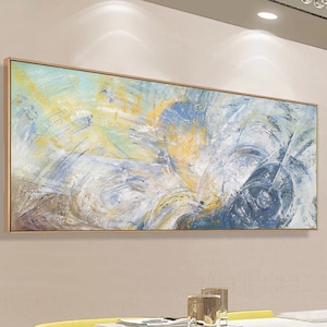 May include: Abstract painting with swirling blue, white, and yellow colors. The painting is framed in a thin gold frame and is hanging on a light beige wall above a dining table.