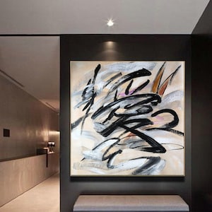 Puede incluir: Pintura abstracta con pinceladas negras, blancas y grises sobre un lienzo beige. La pintura está enmarcada en un marco dorado y cuelga de una pared negra.