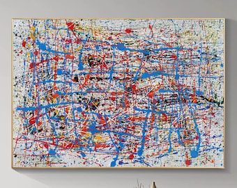 May include: Abstract expressionist painting with a gold frame. The artwork features a white background covered in a chaotic arrangement of colorful paint splatters and lines in red, blue, black, and yellow. The painting is displayed above a wooden table.
