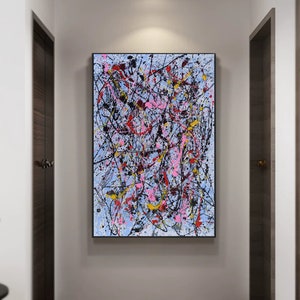 Puede incluir: Pintura abstracta con salpicaduras de pintura negra, blanca, rosa, roja y amarilla sobre un fondo azul claro. La pintura est&aacute; enmarcada en negro y colgada en una pared blanca.