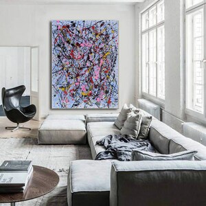 Puede incluir: Pintura abstracta con salpicaduras de pintura negra, rosa, amarilla y roja sobre un fondo azul claro. La pintura est&aacute; colgada en una pared blanca en una sala de estar moderna con un sof&aacute; seccional gris y una mesa de caf&eacute;.