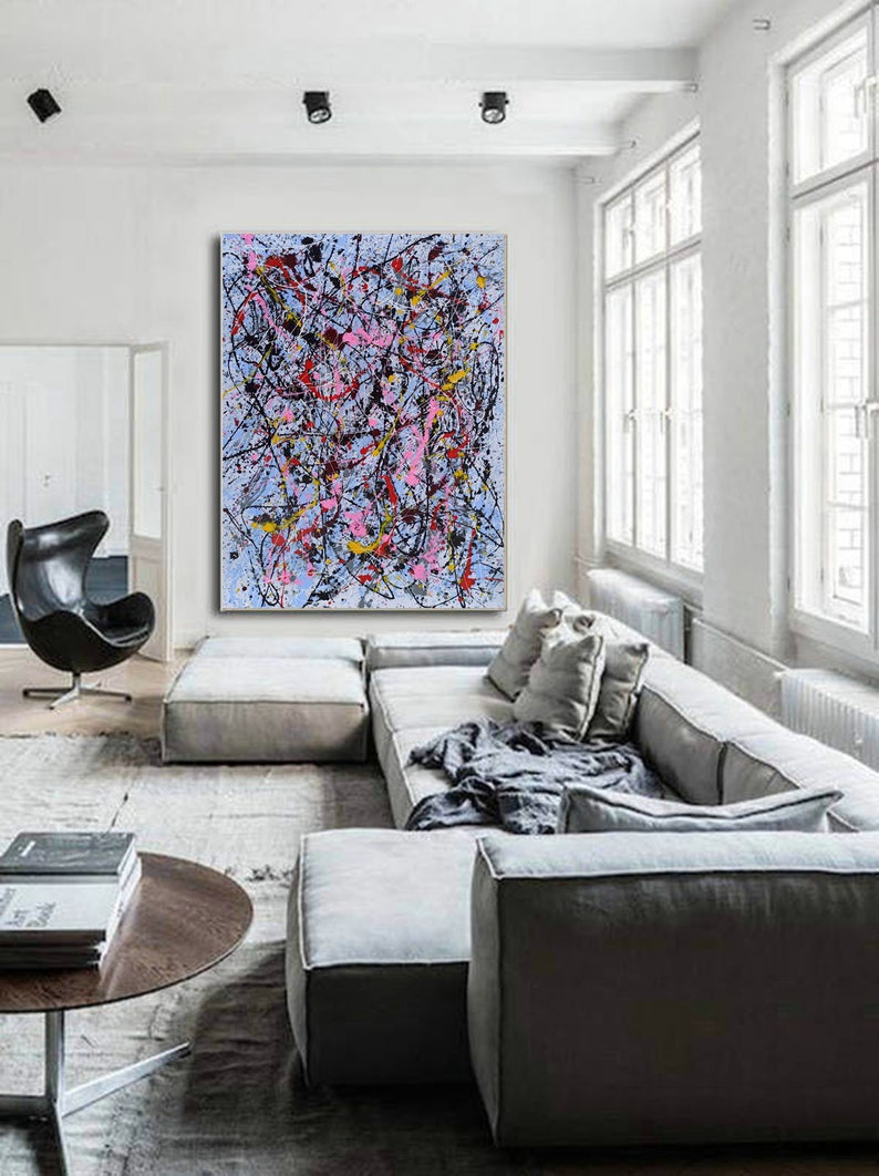 Puede incluir: Pintura abstracta con salpicaduras de pintura negra, rosa, amarilla y roja sobre un fondo azul claro. La pintura est&aacute; colgada en una pared blanca en una sala de estar moderna con un sof&aacute; seccional gris y una mesa de caf&eacute;.
