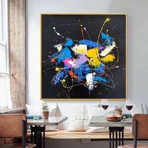 May include: Abstract painting with a black background and colorful splashes of blue, yellow, pink, orange, and white paint. The painting is framed in a gold frame.