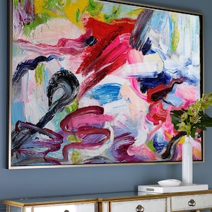 Puede incluir: Pintura abstracta con trazos audaces de pintura rosa, roja, azul, negra y blanca. La pintura está enmarcada en un marco dorado y cuelga de una pared azul.