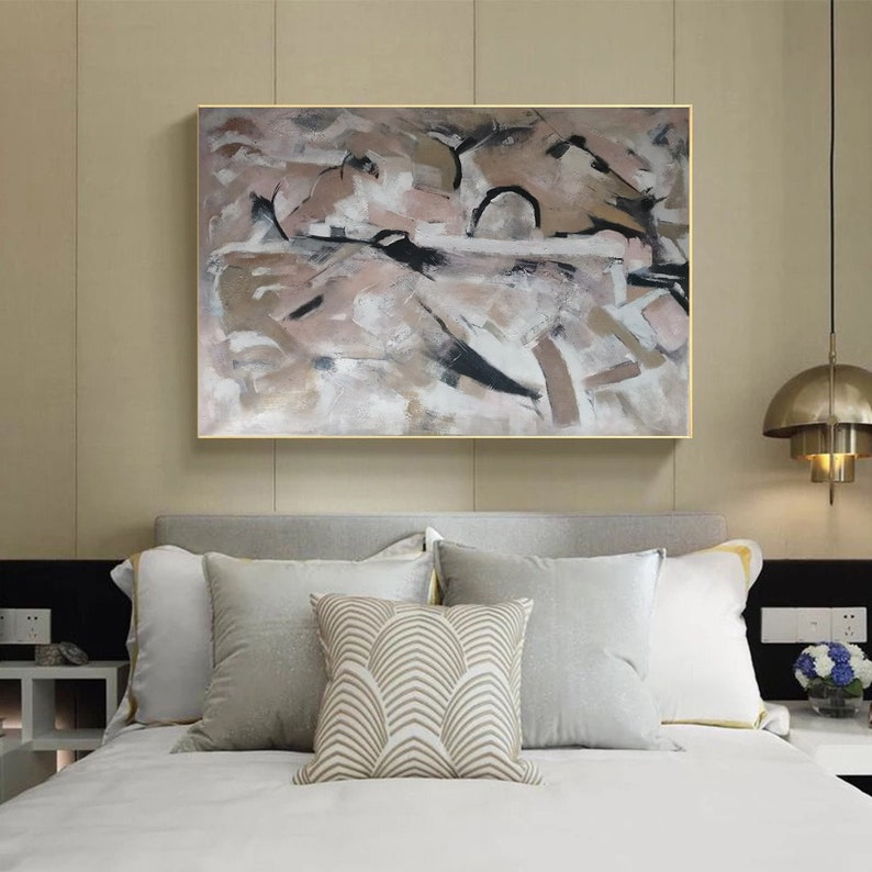 May include: Abstract painting with brown, black, and white brushstrokes on a canvas. The painting is framed in a gold frame and is hanging on a wall above a bed with white bedding and pillows.