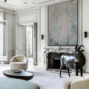May include: A modern living room interior with a white fireplace and a black and white abstract painting above it. A black metal sculpture of a goat stands in front of the fireplace. A white armchair and a round coffee table are in the foreground.