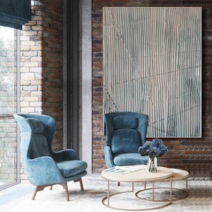 May include: Two blue velvet armchairs with a wooden frame, a round wooden coffee table with a glass vase of blue flowers, and a large abstract painting with white and blue stripes on a brick wall.