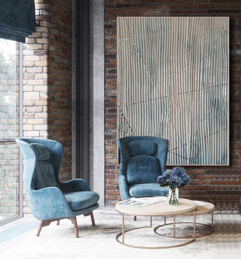 May include: Two blue velvet armchairs with a wooden frame, a round wooden coffee table with a glass vase of blue flowers, and a large abstract painting with white and blue stripes on a brick wall.