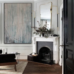 May include: A white fireplace with a black fire grate and a black door. A black leather basket with logs sits in front of the fireplace. A gold-colored wall sconce with a black cord is mounted on the wall above the fireplace. A large abstract painting with blue and white stripes hangs on the wall to the left of the fireplace. A black leather couch sits in the foreground.