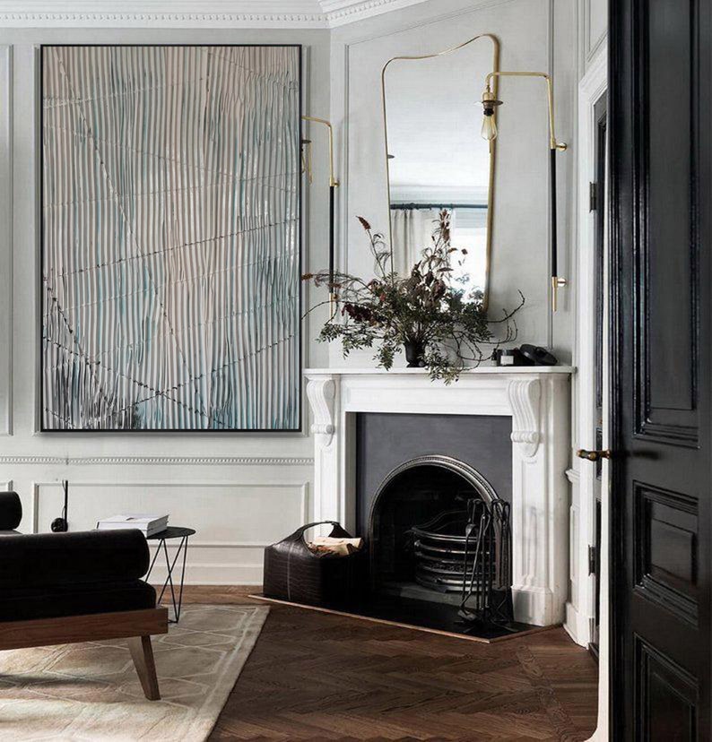 May include: A white fireplace with a black fire grate and a black door. A black leather basket with logs sits in front of the fireplace. A gold-colored wall sconce with a black cord is mounted on the wall above the fireplace. A large abstract painting with blue and white stripes hangs on the wall to the left of the fireplace. A black leather couch sits in the foreground.