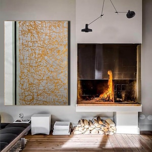 K&ouml;nnte beinhalten: Ein modernes Wohnzimmer mit einem Kamin und einem gro&szlig;en abstrakten Gem&auml;lde mit goldenen Linien auf wei&szlig;em Hintergrund. Das Gem&auml;lde h&auml;ngt an einer wei&szlig;en Wand. Der Kamin ist aus Ziegeln gebaut und hat einen schwarzen Metallrahmen. Vor dem Kamin liegt ein Stapel Brennholz.